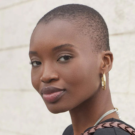 Woman with a short haircut wearing gold hoop earrings and a necklace against a neutral background