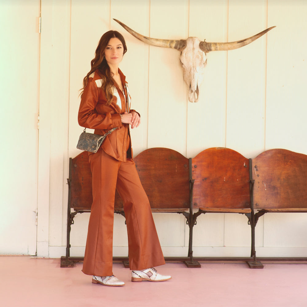 Woman in a brown outfit standing in front of wooden benches and a longhorn skull on a white wall.