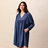A woman wearing a loose, oversized blue denim tunic dress with a V-neck, smocked chest detail, and long sleeves, standing against a textured beige wall.