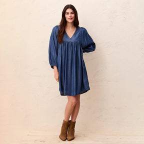 A woman modeling a blue denim-like mini dress with a V-neck, gathered waist, and voluminous elbow-length sleeves, paired with brown ankle boots, standing against a textured cream wall.