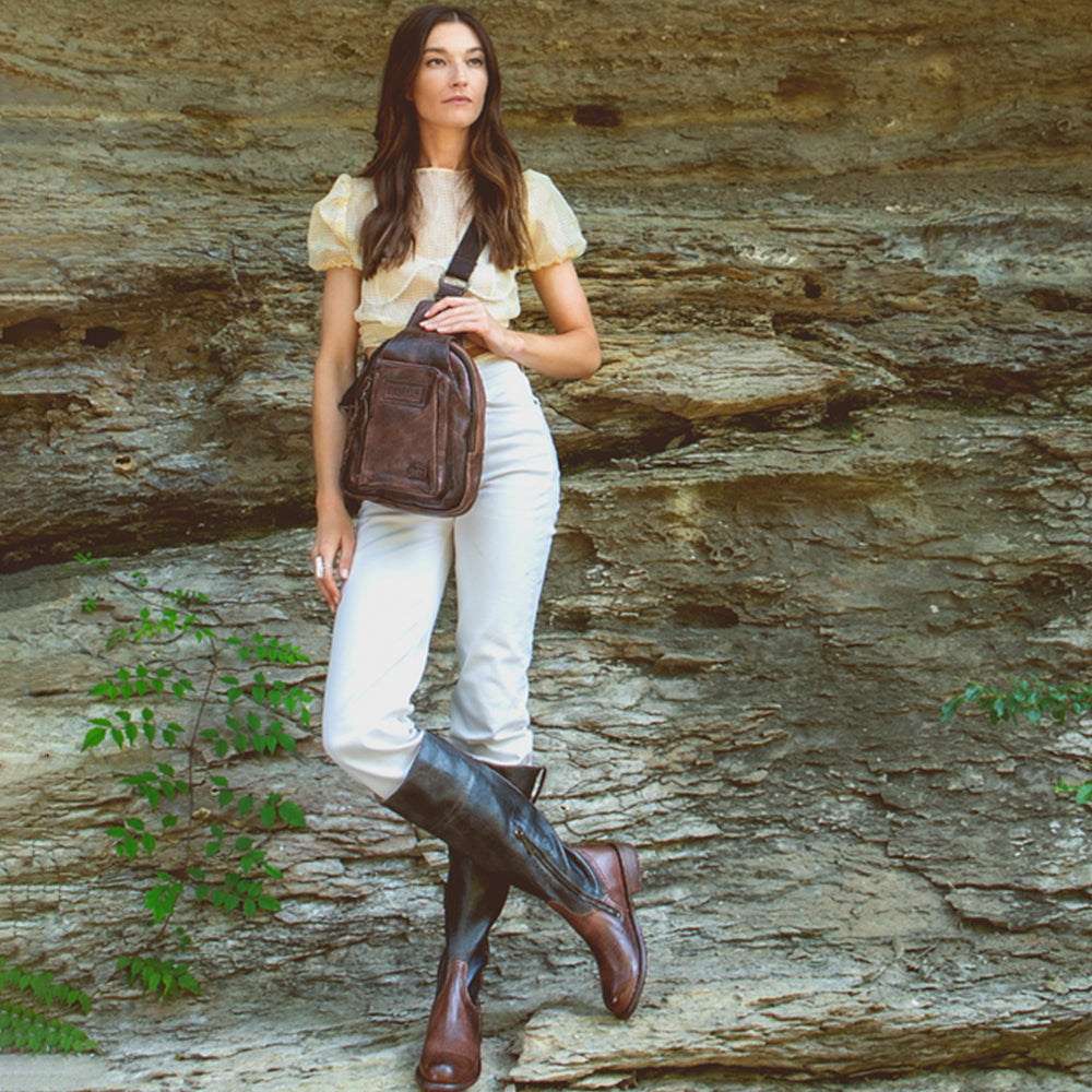 Woman standing outdoors against a rocky background holding a brown leather bag.
