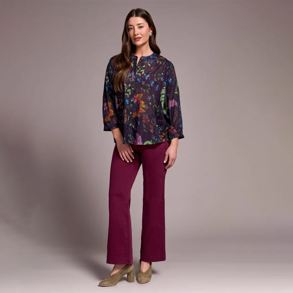 Woman wearing a colorful floral blouse and purple pants against a plain background
