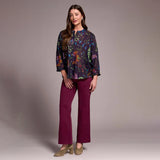 Woman wearing a colorful floral blouse and purple pants against a plain background
