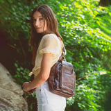 Woman holding a brown leather bag in a natural setting with greenery