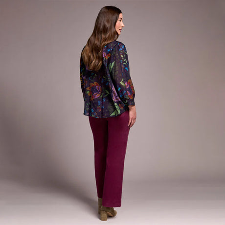 Woman wearing a colorful floral blouse and purple pants against a plain background