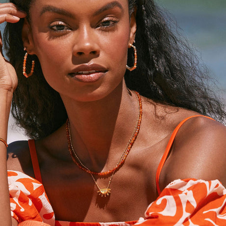 Woman wearing an orange and white patterned top with a necklace, against a blurred natural background.