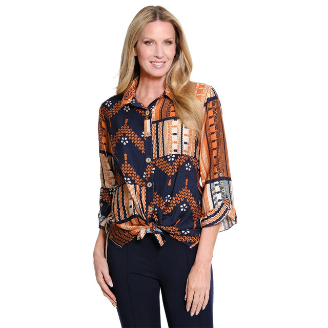 A woman modeling a long-sleeve button-up blouse with a geometric tribal-inspired print in navy blue, orange, and beige, paired with dark pants against a plain white background.