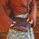 Person wearing a brown knitted sweater holding a brown leather bag with a blurred wooden background