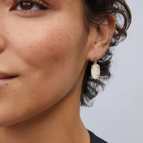 Close-up of a person wearing a gold earring with a light stone on a neutral background