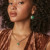 Woman wearing a gold necklace and green earrings with a brown top.