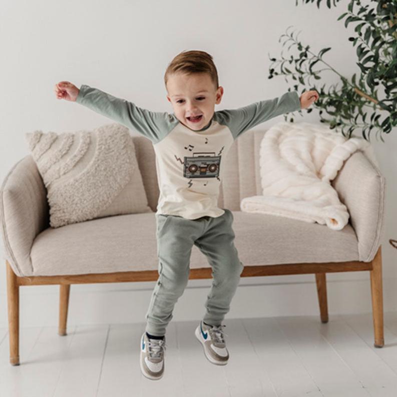 A toddler wearing a long sleeve baseball tee with a boombox graphic, jumping and smiling in a room with a sofa and a plush toy.