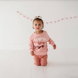 A toddler wearing a pink long-sleeve sweatshirt with a hearts pattern and drop shoulders, posing on a white background.