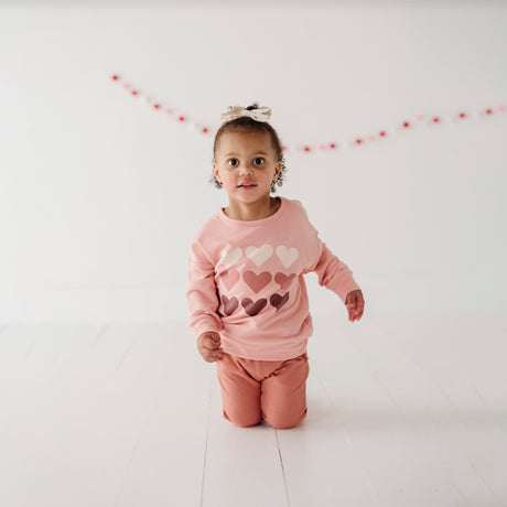 A toddler wearing a pink long-sleeve sweatshirt with a hearts pattern and drop shoulders, posing on a white background.
