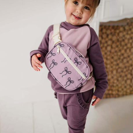 Child wearing a purple outfit with a matching bag, standing against a neutral background.