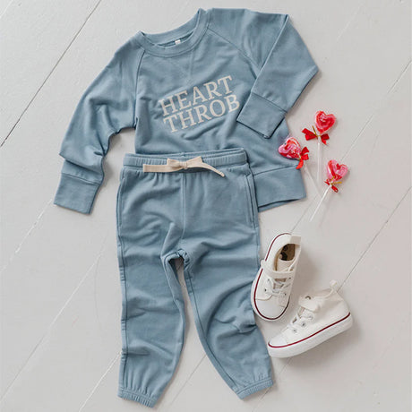Children's blue outfit with 'HEART THROB' text, white sneakers, and decorative flowers on a light wooden floor.