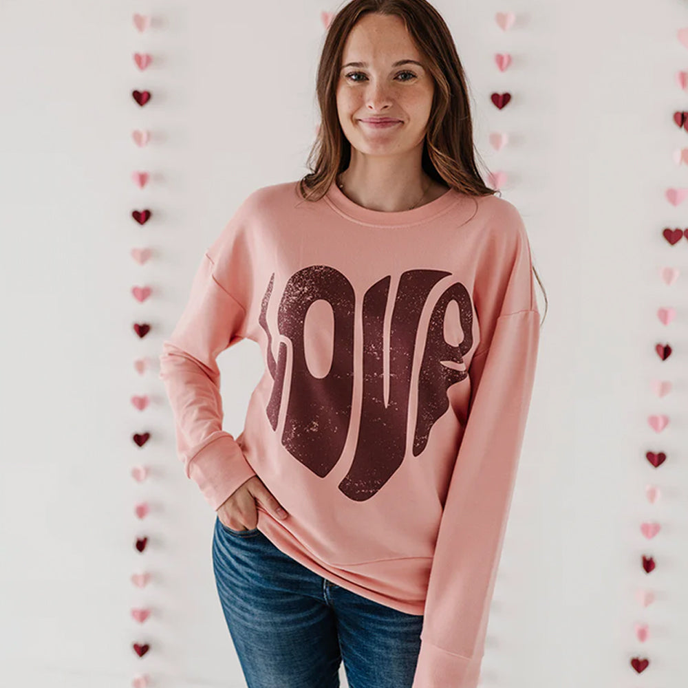 Woman wearing a pink sweatshirt with brown text, standing against a wall with heart decorations.