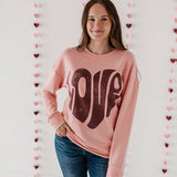 Woman wearing a pink sweatshirt with brown text, standing against a wall with heart decorations.