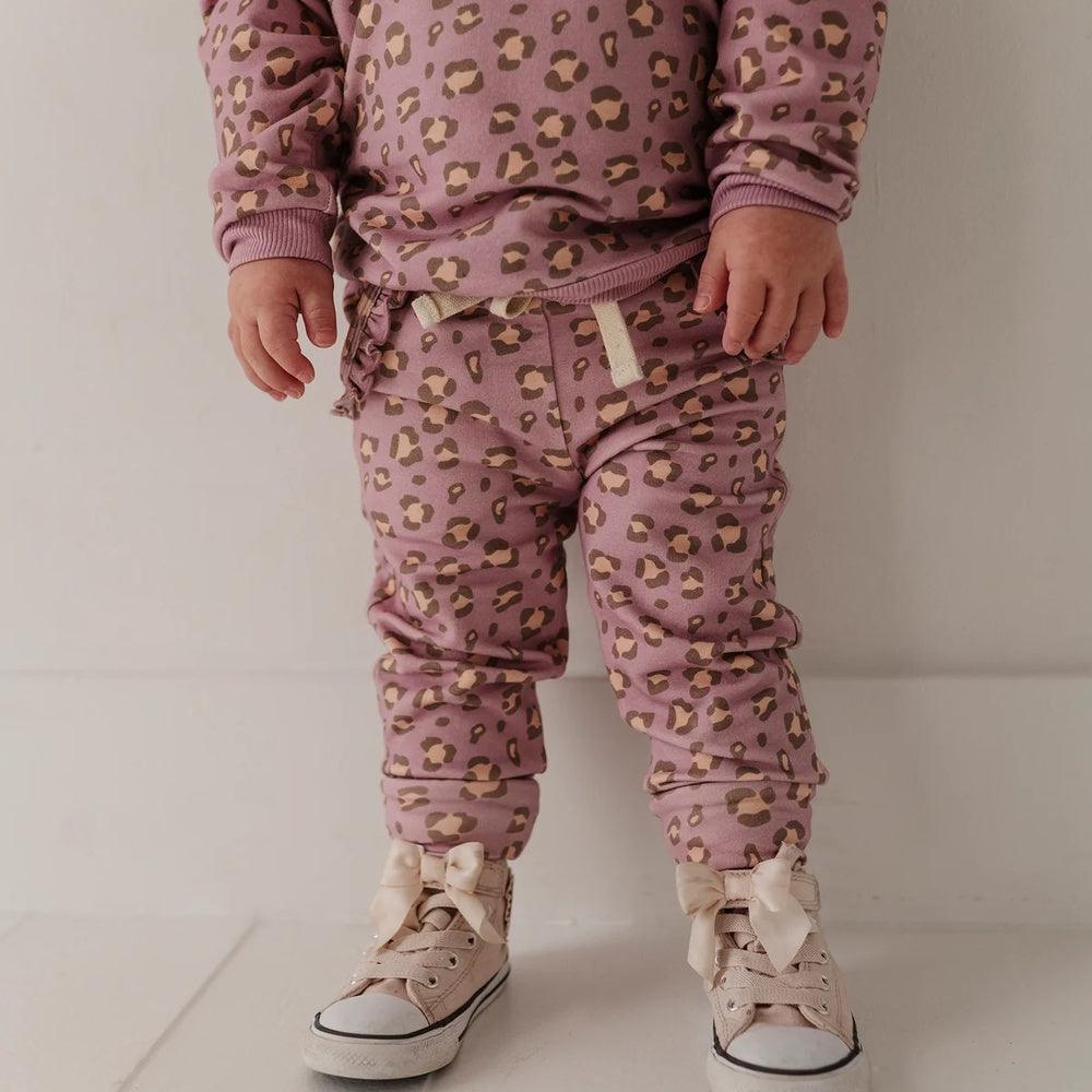 A toddler standing indoors wearing a matching mauve pajama set with a tan and brown leopard print, cream drawstrings, and cream lace-up sneakers with bows.