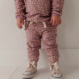 A toddler standing indoors wearing a matching mauve pajama set with a tan and brown leopard print, cream drawstrings, and cream lace-up sneakers with bows.