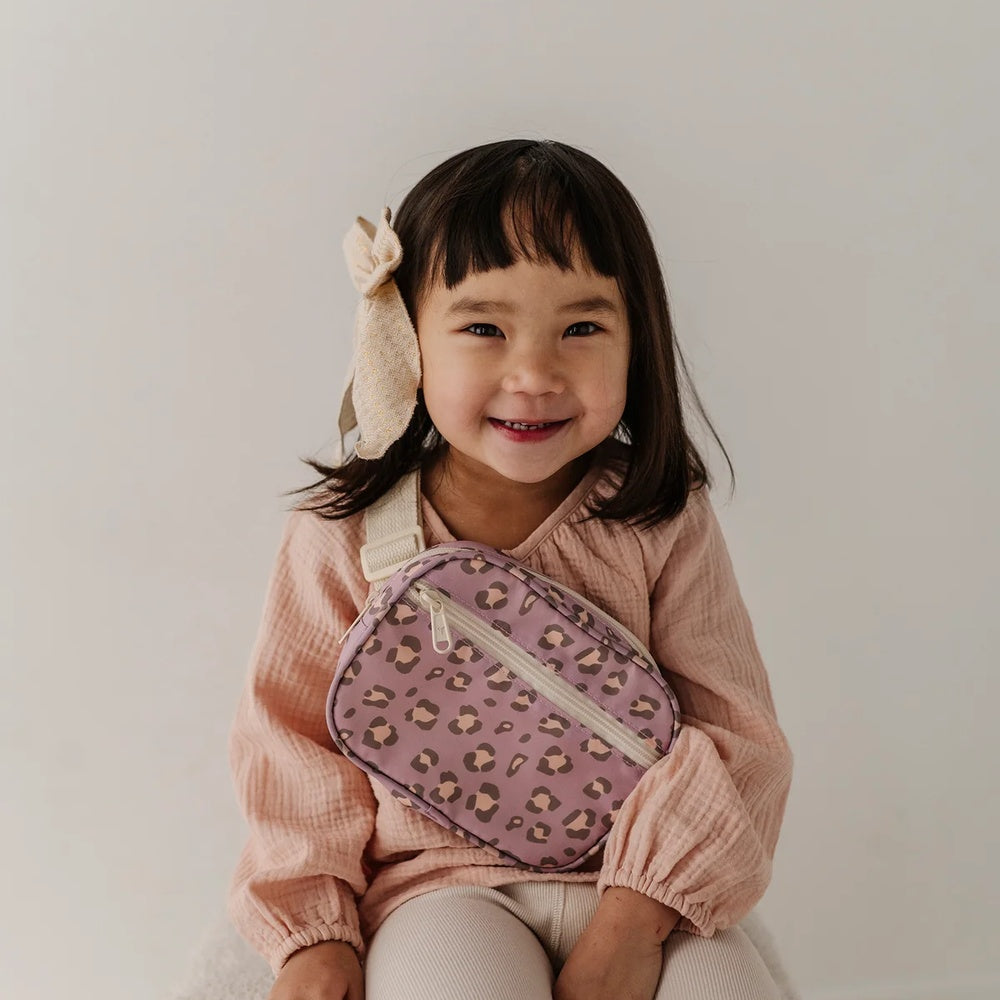 A young girl with a beige bunny hair bow smiles while wearing a pink textured top and holding a pink leopard-print crossbody bag with a white strap.