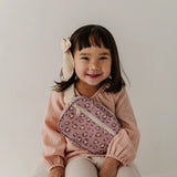 A young girl with a beige bunny hair bow smiles while wearing a pink textured top and holding a pink leopard-print crossbody bag with a white strap.