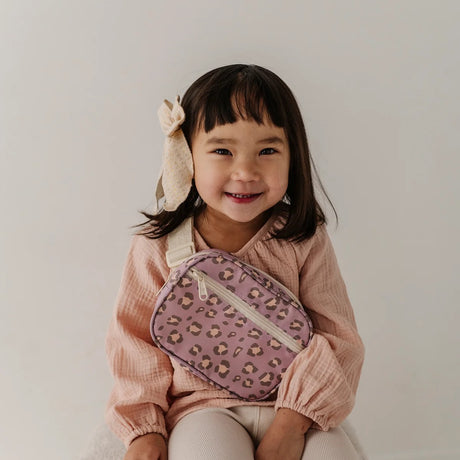 A young girl with a beige bunny hair bow smiles while wearing a pink textured top and holding a pink leopard-print crossbody bag with a white strap.