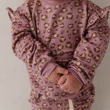 A close-up of a child wearing a dusty mauve pajama set with a brown and beige leopard print pattern, featuring ruffle details on the sleeves and a matching headband.