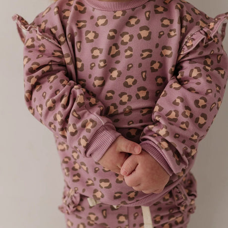 A close-up of a child wearing a dusty mauve pajama set with a brown and beige leopard print pattern, featuring ruffle details on the sleeves and a matching headband.