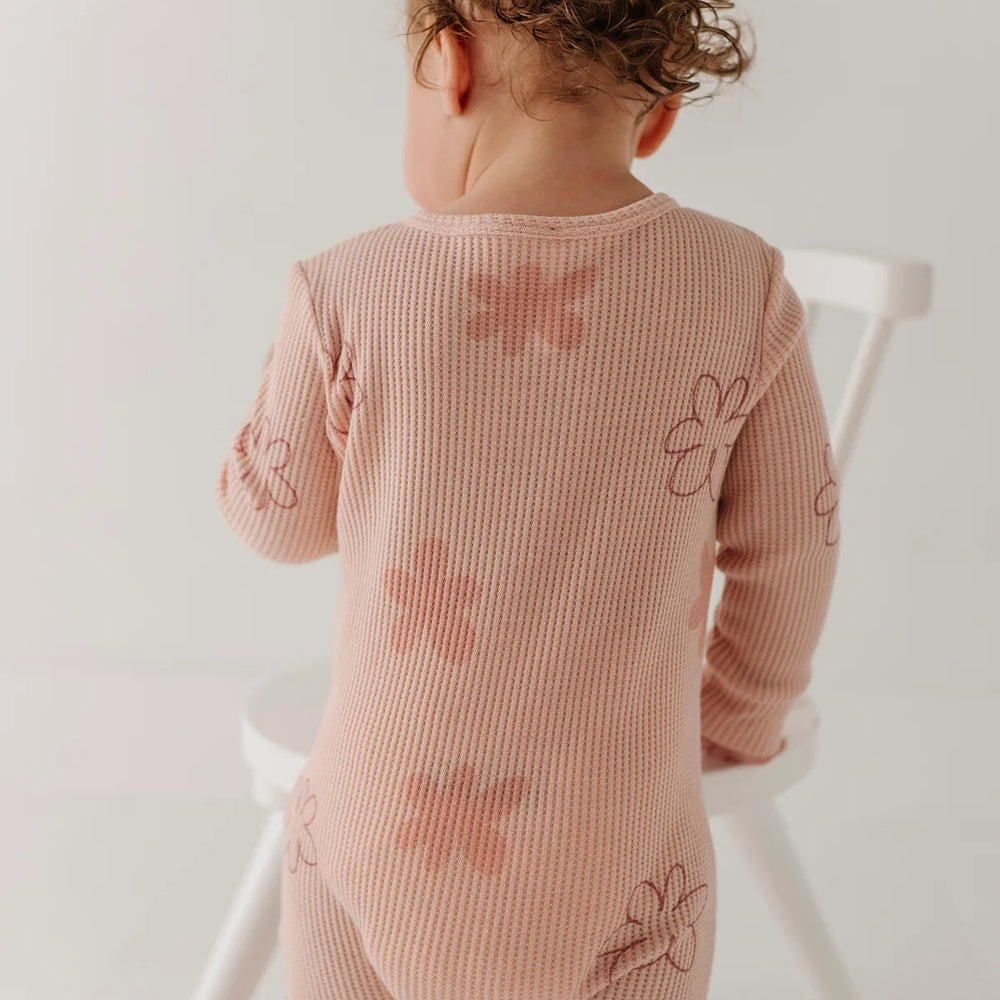 A baby wearing a dusty pink ribbed long-sleeve bodysuit with embroidered flower patterns, photographed from behind while sitting on a white wooden chair against a plain white background.