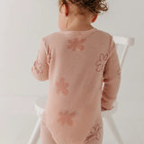 A baby wearing a dusty pink ribbed long-sleeve bodysuit with embroidered flower patterns, photographed from behind while sitting on a white wooden chair against a plain white background.