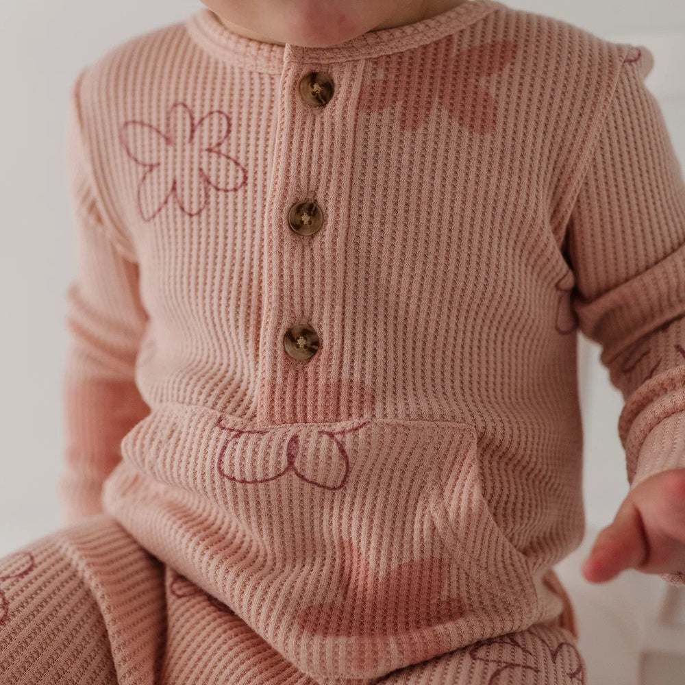 A close-up of a baby wearing a dusty pink ribbed knit romper with brown buttons and embroidered pink floral details.