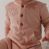 A close-up of a baby wearing a dusty pink ribbed knit romper with brown buttons and embroidered pink floral details.