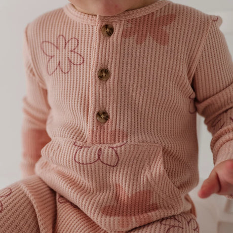 A close-up of a baby wearing a dusty pink ribbed knit romper with brown buttons and embroidered pink floral details.