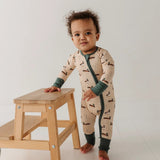 A baby wearing a beige long-sleeve footless sleeper with a green zipper and dark green cuffs, standing next to a small wooden step stool.