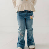 A child wearing blue denim jeans with colorful embroidered patches, paired with a cream-colored ruffled top and white shoes, shown from the waist down against a light studio background.