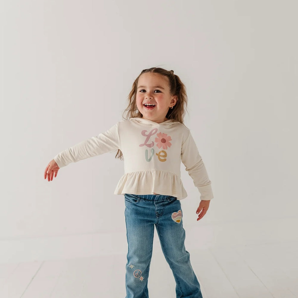 A young girl with light brown hair in a ponytail is smiling and posing in a studio. She is wearing a cream-colored long-sleeve top with a 'Love' graphic and a ruffled hem, paired with blue jeans featuring heart and star patches.