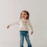 A young girl with light brown hair in a ponytail is smiling and posing in a studio. She is wearing a cream-colored long-sleeve top with a 'Love' graphic and a ruffled hem, paired with blue jeans featuring heart and star patches.