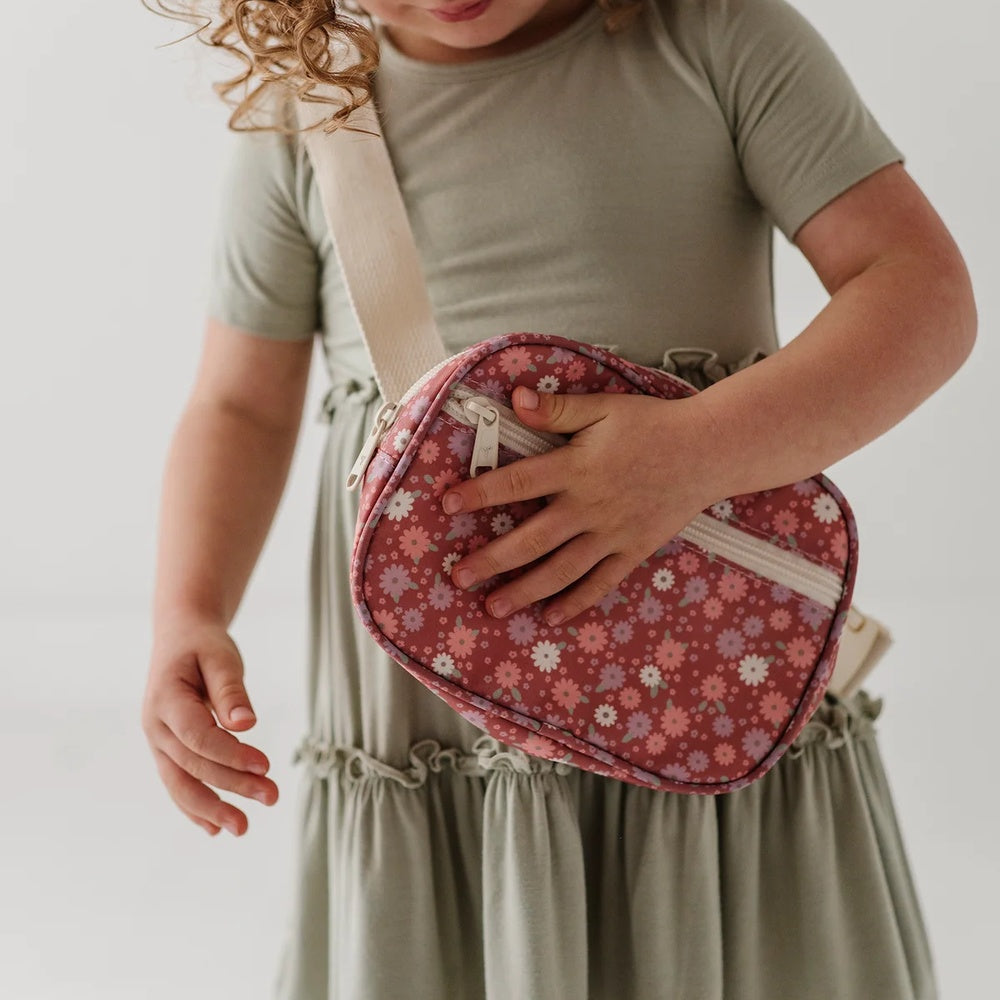 A child wearing an olive green dress holds a small, rounded crossbody bag with a pink floral pattern and a beige strap against a plain white background.
