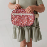 A child wearing an olive green dress holds a pink floral crossbody bag with a beige buckle strap against a plain light background.