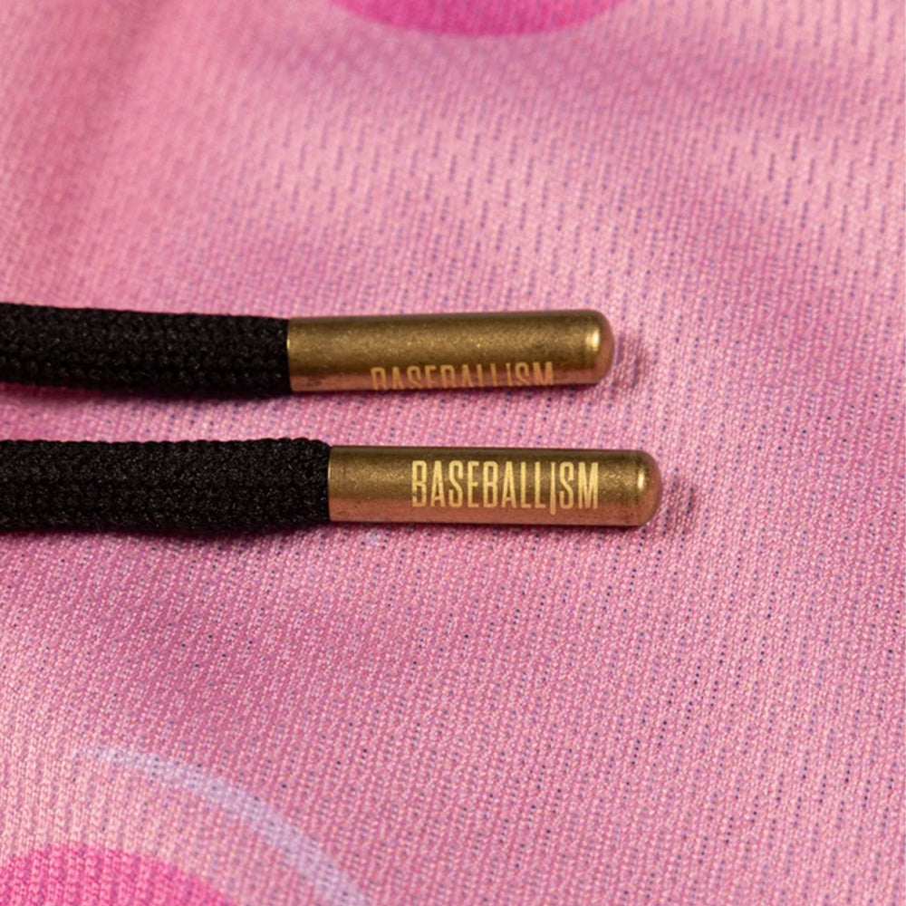 Close-up of two black braided cord ends with metallic gold cylindrical caps printed with the word 'BASEBALLISM', resting on a pink textured fabric background.