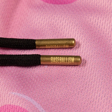Close-up of two black braided cord ends with metallic gold cylindrical caps printed with the word 'BASEBALLISM', resting on a pink textured fabric background.