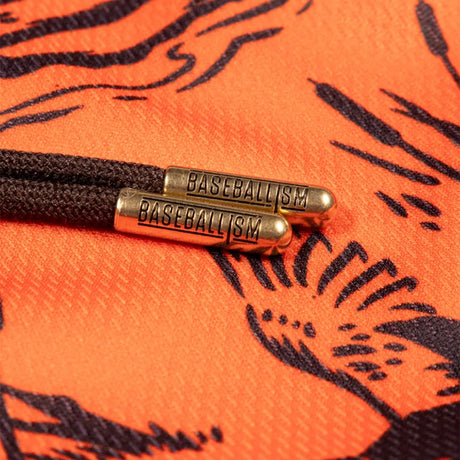Close-up of a dark brown braided cord with two metallic gold-toned sliders engraved with the word 'BASEBALLISM', resting on an orange fabric background with a black graphic print.