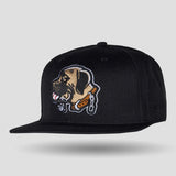 A black flat-brim snapback cap featuring a large embroidered patch of a dog's head on the front. The patch is tan and brown with white and black outlines, and the cap itself is shown against a light gray background.