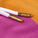 Close-up of white braided drawstring cords with gold metal tips engraved with the word 'BASEBALLISM', resting on a textured fabric background split between orange and magenta.
