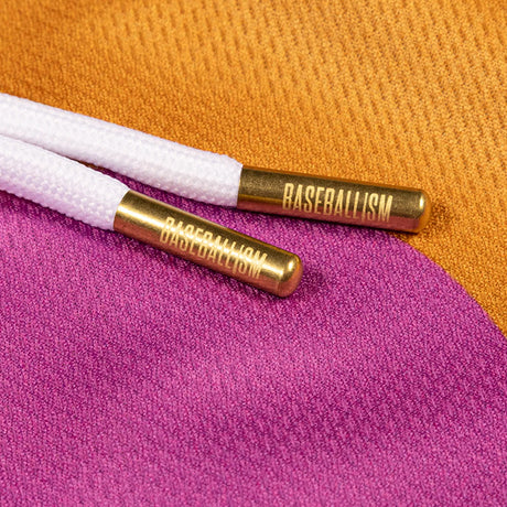 Close-up of white braided drawstring cords with gold metal tips engraved with the word 'BASEBALLISM', resting on a textured fabric background split between orange and magenta.