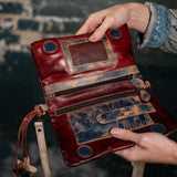 A close-up of a person holding an open distressed leather clutch or wallet. The item is made of deep red and dark blue leather with heavily weathered tan patches, showing interior compartments, zippers, and circular cutouts.