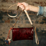 A hand holds a small, dark red leather shoulder bag with a cream-colored strap and leather charms against a rustic, blurred brick wall background.