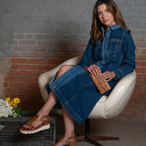 Woman in a denim dress sitting on a white chair against a brick wall.