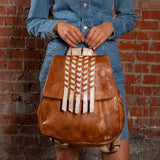 Person holding a brown leather backpack with white fringe against a brick wall.
