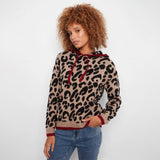 A woman with curly brown hair wearing a beige and black leopard print hoodie with dark red trim and drawstrings, paired with blue jeans, against a plain white background.